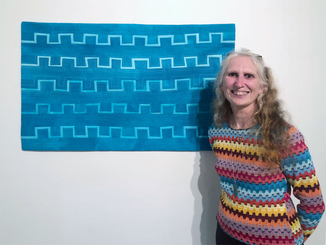 Red headed artist wearing stripey top is smiling next to her aqua-hued textile painting
